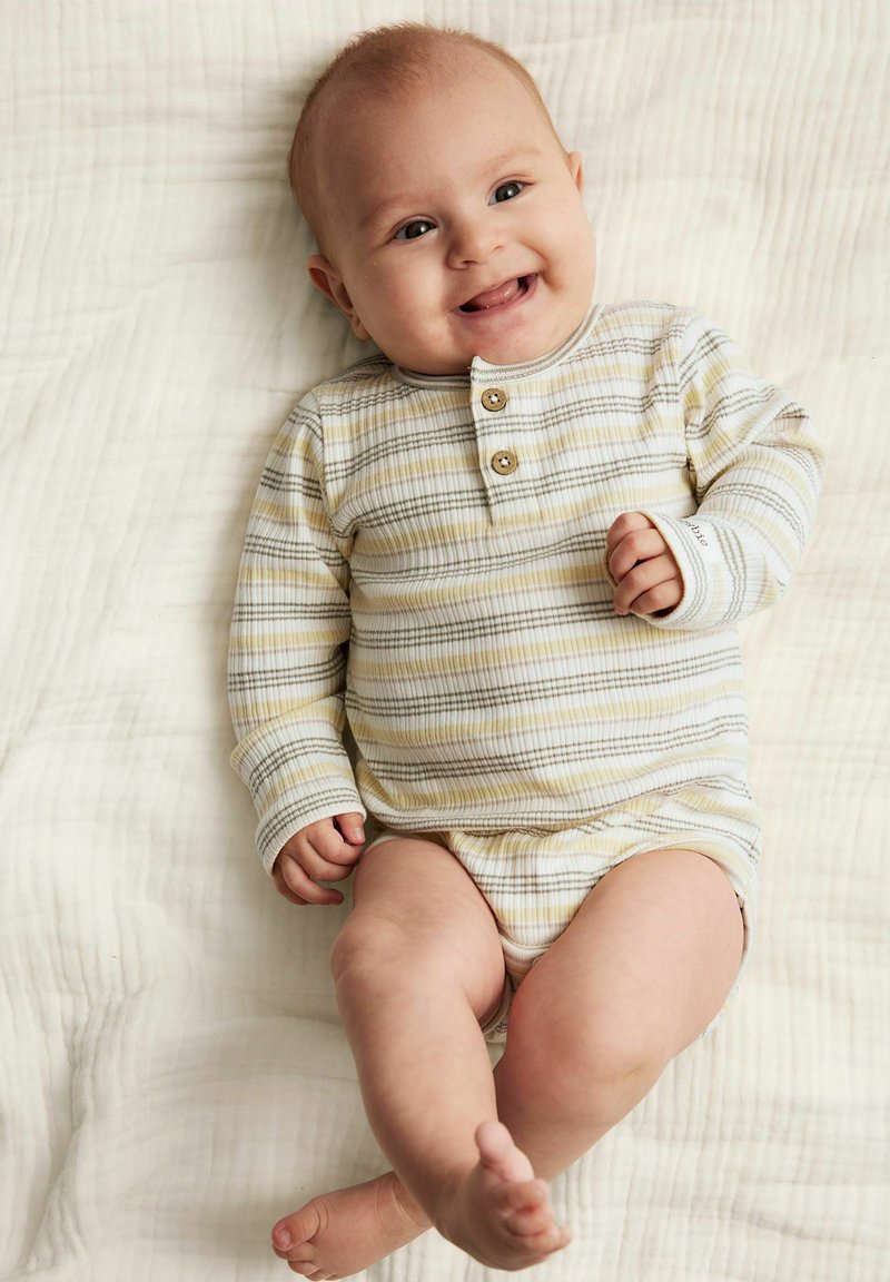 Un body à manches longues, rayé jaune clair et gris, avec des boutons en bois. Assis sur une couverture blanche texturée. L'expression joyeuse du bébé est évidente.