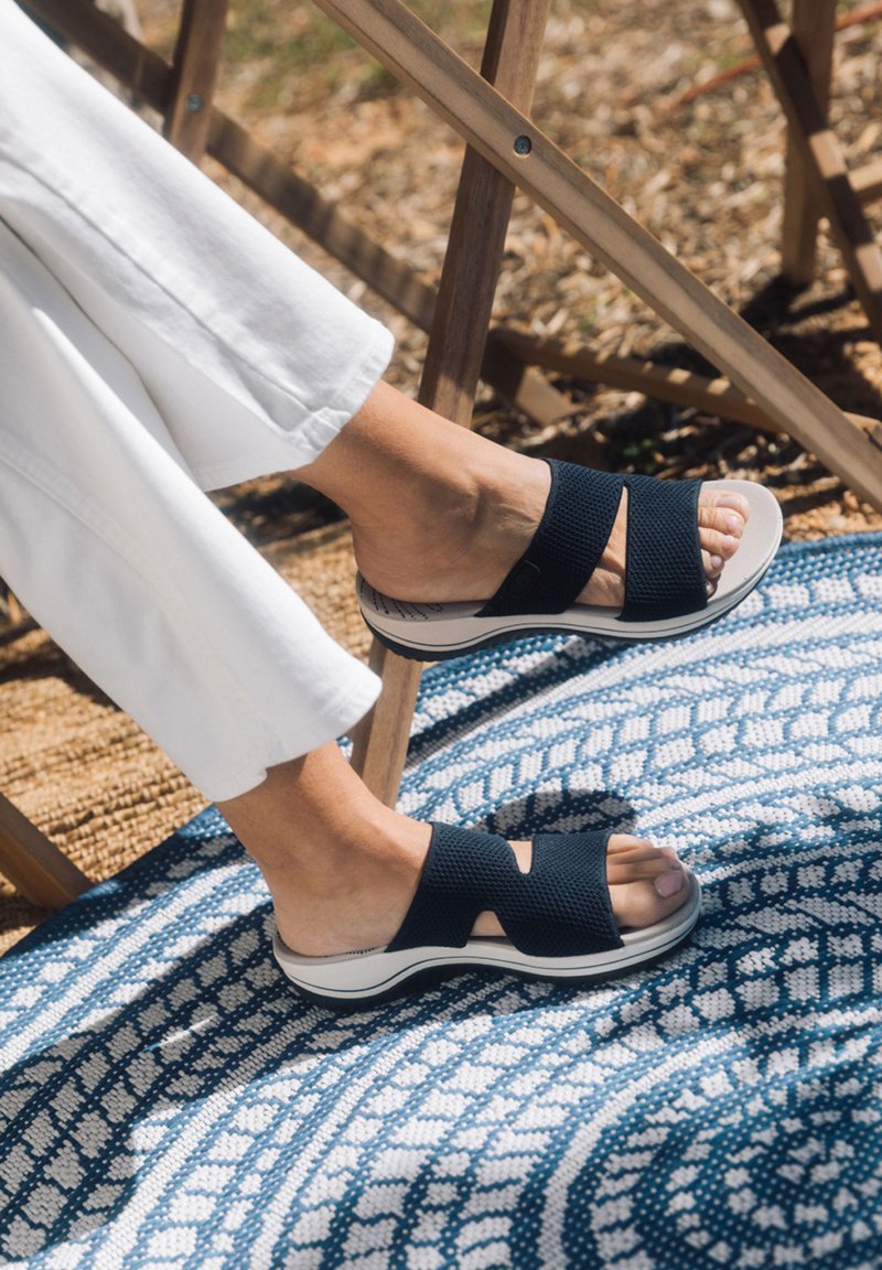Schwarze Mesh-Sandalen mit Kreuzriemen, ausgestattet mit einer gepolsterten weißen Sohle und einem strukturierten Fußbett, auf einem gemusterten blauen Teppich.
