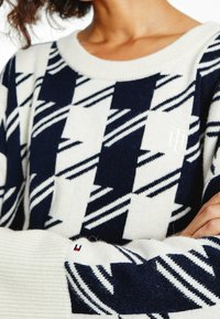 Marineblauer und weißer Strickpullover mit geometrischem Muster, geripptem Kragen und Logodetail am Ärmel. Weicher Stoff und lockere Passform.