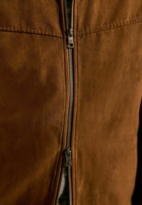 Nahaufnahme einer braunen Wildlederjacke mit einem doppelt ausgezippbaren Reißverschluss, die teilweise geöffnet ist, sodass das Innenfutter sichtbar wird.