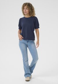 Navy blue short-sleeve top with puffed sleeves, paired with light blue flared jeans and beige sneakers, set against a plain white background.