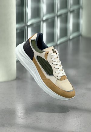 Zapatilla beige, marrón claro y verde con suela y cordones blancos, flotando sobre un suelo de cemento gris con fondo de pared de bloques de vidrio.