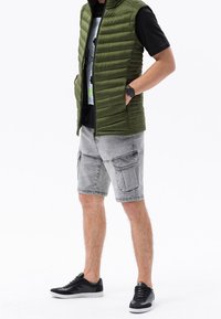 Zielona kamizelka puchowa noszona na czarnym T-shircie, zestawiona z jasnoszarymi szortami cargo i czarnymi tenisówkami. Charakterystyczne cechy to zamek błyskawiczny i boczne kieszenie.