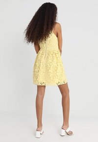 Keltainen pitsimekko, jossa on levenevä hame ja takana vetoketju, yhdistettynä valkoisiin paksupohjaisiin sandaaleihin. Teksturoitu pitsikuvio.