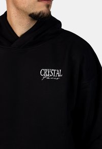 Czarna bluza z kapturem o luźnym kroju, z dużą kieszenią z przodu, kapturem na sznurki oraz białym haftowanym logo "CRYSTAL Paris" na klatce piersiowej.