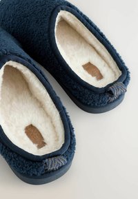 Mörkblå tofflor med en fluffig yta, krämfärgad fleecefoder och läderetikett. Har en texturerad ögla vid hälen.