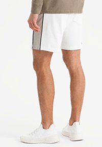 Pantaloni corti bianchi con strisce nere laterali, realizzati in cotone, con una tasca sul retro e una texture liscia. Abbinati a sneakers bianche.