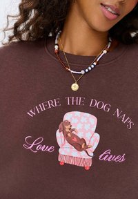 Hnědá mikina s růžovým grafickým motivem spícího psa na vzorované židli. Text zní "KDE PES SPÍ" v hravém písmu.