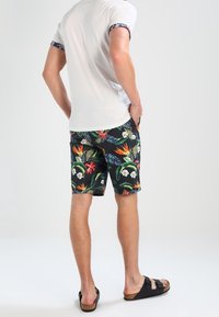 Uomo in piedi con le mani nelle tasche, indossa una t-shirt bianca, pantaloni corti neri a fiori e sandali neri su uno sfondo chiaro e semplice.
