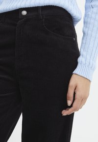 Zwarte corduroy broek met verticale ribbels, voorzien van een zak aan de voorkant en een zilveren knoopdetail bij de taille, gecombineerd met een lichtblauwe gebreide trui.