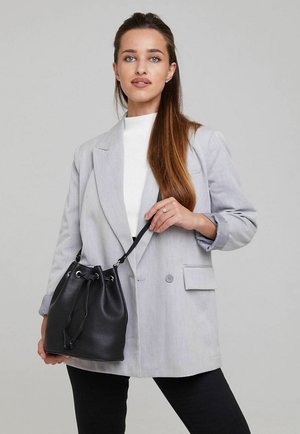 Frau mit langen braunen Haaren, die einen hellgrauen Blazer und ein weißes Top trägt, hält eine schwarze Leder-Bucket-Bag mit Kordelverschluss.