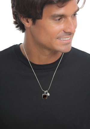 Mann in schwarzem Hemd trägt eine silberne Kette mit quadratischem dunklen Stein und kleinem ovalem Anhänger, blickt nach rechts und lächelt leicht.