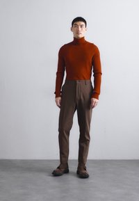 Roestkleurige geribbelde coltrui, gecombineerd met bruine op maat gemaakte broeken. Model staat voor een neutrale achtergrond en draagt donkere schoenen.