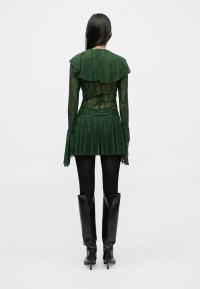 Πράσινο φόρεμα με δαντέλα, με λουλουδάτο λαιμόκοψη, μανίκια καμπάνα, πλισέ φούστα, και συνδυασμένο με εφαρμοστά μαύρα μπότες πάνω από το γόνατο.