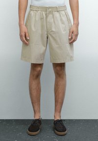 Beige katoenen shorts met een elastische tailleband, twee voorzakken en een relaxte pasvorm, gecombineerd met donkere schoenen.