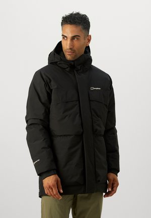 Muž nosiaci čiernu zimnú bundu Berghaus s kapucňou z materiálu Gore-Tex a zelené nohavice, pozerajúci sa do strany na jednoduchom pozadí.