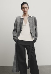 Cardigan en laine gris porté sur un top blanc à col en V, associé à un pantalon noir avec une ceinture. Texture lisse et coupe ajustée tout au long.
