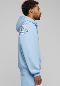Light blue hoodie with a front zipper, side pockets, and a stylized design on the left sleeve. Made from soft, textured fabric.