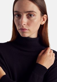 Schwarzer Rollkragenpullover aus weichem Strickstoff, mit geripptem Kragen und Bündchen, getragen von einem Model, das seinen Arm hält. Glatte Textur, minimalistisches Design.