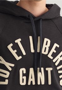 Šedá mikina s kapucí ve tmavé barvě s krémovým logem se zvýšeným písmem, stahovacími šňůrkami v tmavším odstínu a měkkou, texturovanou tkaninou.