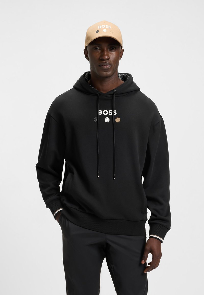 Felpa nera con logo "BOSS" in bianco e tre colori di accento. Presenta una coulisse e polsini a coste. Abbinata a un cappellino beige.
