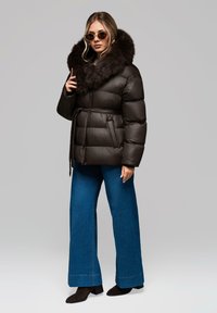 Giacca nera a compressione con colletto in finta pelliccia, dotata di vita da annodare, cuciture orizzontali e tasche laterali. Il modello l'ha abbinata a pantaloni larghi blu.