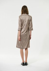 Braun gemustertes Kleid mit Dreiviertelärmeln, einem Kragen und gerader Schnittführung, das geometrische Formen in gedeckten Farben zeigt. Schwarze Schuhe getragen.