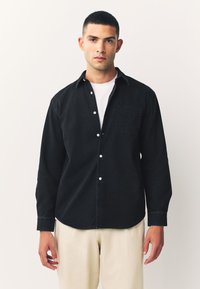 Schwarzes Hemd mit Knopfleiste und schmaler Passform, ausgestattet mit einer Brusttasche, einem Button-Down-Kragen und weißen Knöpfen, kombiniert mit hellbeigen Hosen.