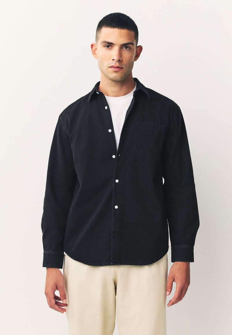 Schwarzes Hemd mit Knopfleiste und schmaler Passform, ausgestattet mit einer Brusttasche, einem Button-Down-Kragen und weißen Knöpfen, kombiniert mit hellbeigen Hosen.