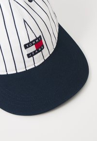 Striped cap featuring a white crown, dark blue brim, and a stitched ‘TOMMY JEANS’ label. Material appears cotton with a textured surface.