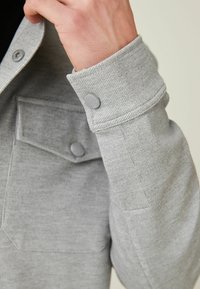 Sudadera gris con un acabado texturizado, que presenta un puño abotonado, un bolsillo en el pecho y detalles de costura en contraste.