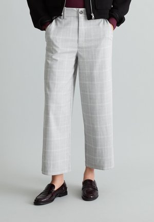 Partie basse du corps portant un pantalon court à carreaux gris clair avec poches latérales et mocassins en cuir marron foncé sur un fond gris uni.