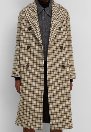 Person iført en beige og grønternet dobbeltradet lang frakke over en mørkegrå lynlåstrøje og sorte bukser.