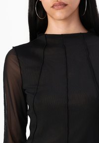 Črn dolg rokav s teksturirano, prozorno plastjo. Ima vertikalne šive in visok ovratnik. Lahka, oprijeta zasnova.