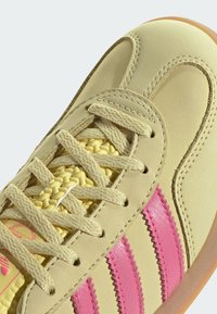 Tenisice svijetložute kožne gornje površine, s ružičastim kožnim trakama, pletenim žutim vezicama i svijetlosmešanim gumeni potplat; glatka tekstura i valoviti akcenti.