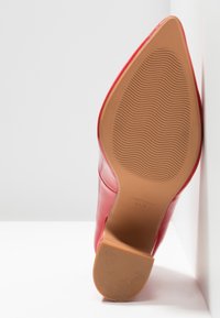 Chaussure à talon haut en cuir verni rouge avec un bout pointu, une surface lisse et une semelle beige texturée présentant un motif ondulé.
