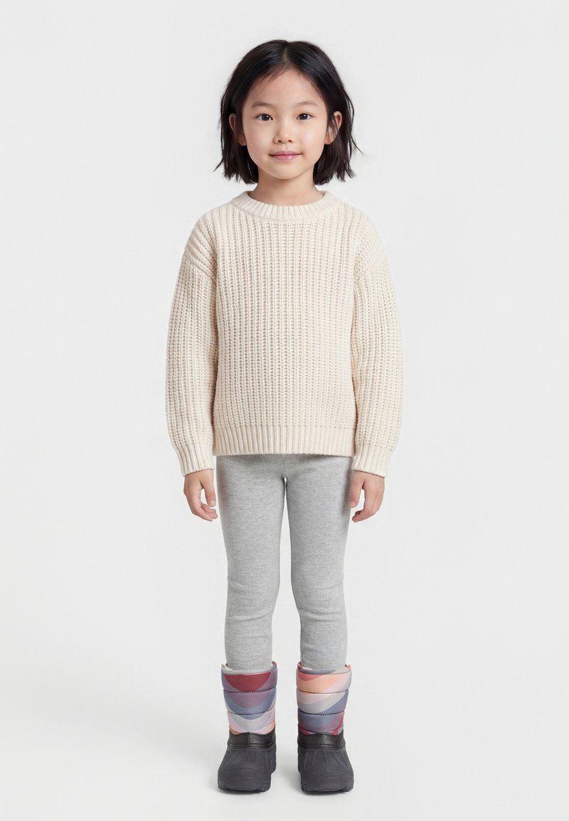 Νεαρή κοπέλα που στέκεται με κρεμ πλεκτό πουλόβερ, ανοιχτό γκρι κολάν και πολύχρωμα επενδυμένα χειμωνιάτικα μποτάκια, μπροστά σε λευκό φόντο.