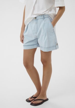 Osoba nosi svetlomodré riflové šortky s vyhrnutými lemami, biele voľné tričko a čierne žabky, stojí na bielom pozadí.