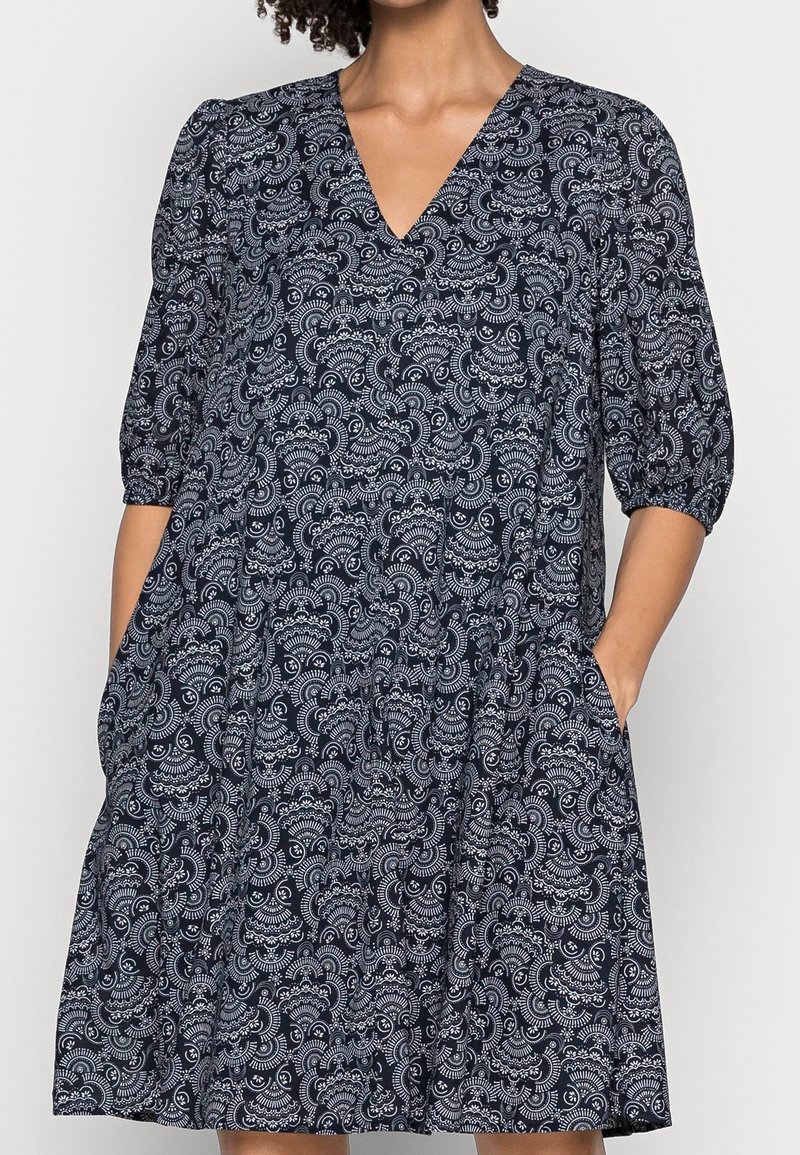 Femme portant une robe bleu marine avec des motifs floraux blancs complexes, manches trois-quarts, encolure en V, et mains dans les poches latérales.