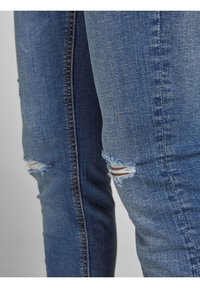 Primer plano de unos vaqueros de mezclilla azul con pequeños desgastes en las rodillas, mostrando la textura de la tela y los detalles de la costura.