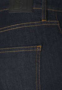 Tmavé džíny vyrobené z denimu s kontrastním oranžovým šitím, hladkým povrchem a černou koženou nášivkou s vyraženým designem na pase.