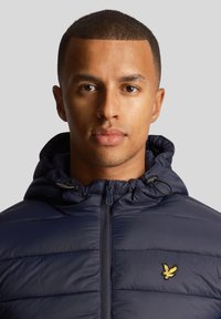 Navy puffer jacket with a zippered front, drawstring hood, and quilted texture. Features a small embroidered logo in yellow on the chest.