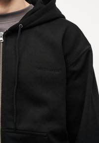 Άτομο που φοράει μαύρο φούτερ με φερμουάρ και κεντημένο το «Last Resort AB» στο στήθος, πάνω από γκρι μπλουζάκι με στρογγυλή λαιμόκοψη.