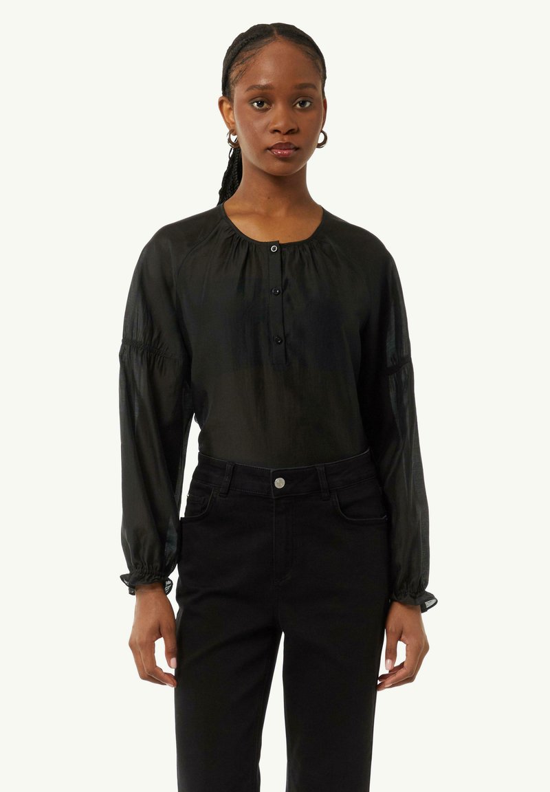 Schwarze Bluse mit langen Ärmeln, rundem Ausschnitt, Knopfleiste, strukturiertem Stoff und gerafften Manschetten, kombiniert mit hochgeschnittenen schwarzen Jeans.