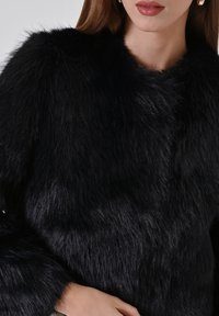 Veste en fausse fourrure noire avec une texture douce et dense, dotée d'un col arrondi et de manches longues. Vue en gros plan mettant en valeur la brillance du matériau.