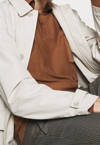 Impermeabile beige chiaro con grandi bottoni, indossato sopra una polo a coste marrone, abbinato a pantaloni in tessuto a quadri. Texture liscia, vestibilità sartoriale.