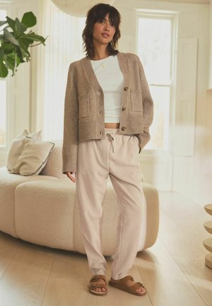 Cardigan beige a maglia con polsini a coste, top bianco aderente e pantaloni a righe chiare, abbinati a sandali marroni con doppie cinghie.