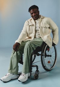 Fehér fleece dzseki, zöld kockás nadrág és piros akcentusokkal ellátott fehér sportcipő. A modell kerekesszékben ül, kék háttér előtt.