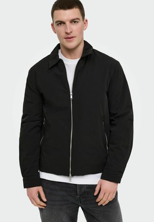 Mladi moški, oblečen v črno jakno z zadrgo čez belo majico in temnosive jeans hlače, stoji pred enostavnim svetlim ozadjem.