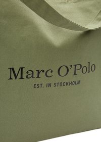 Sac en tissu vert avec "Marc O'Polo EST. À STOCKHOLM" imprimé en noir, montrant un gros plan de la texture et des détails de la sangle.